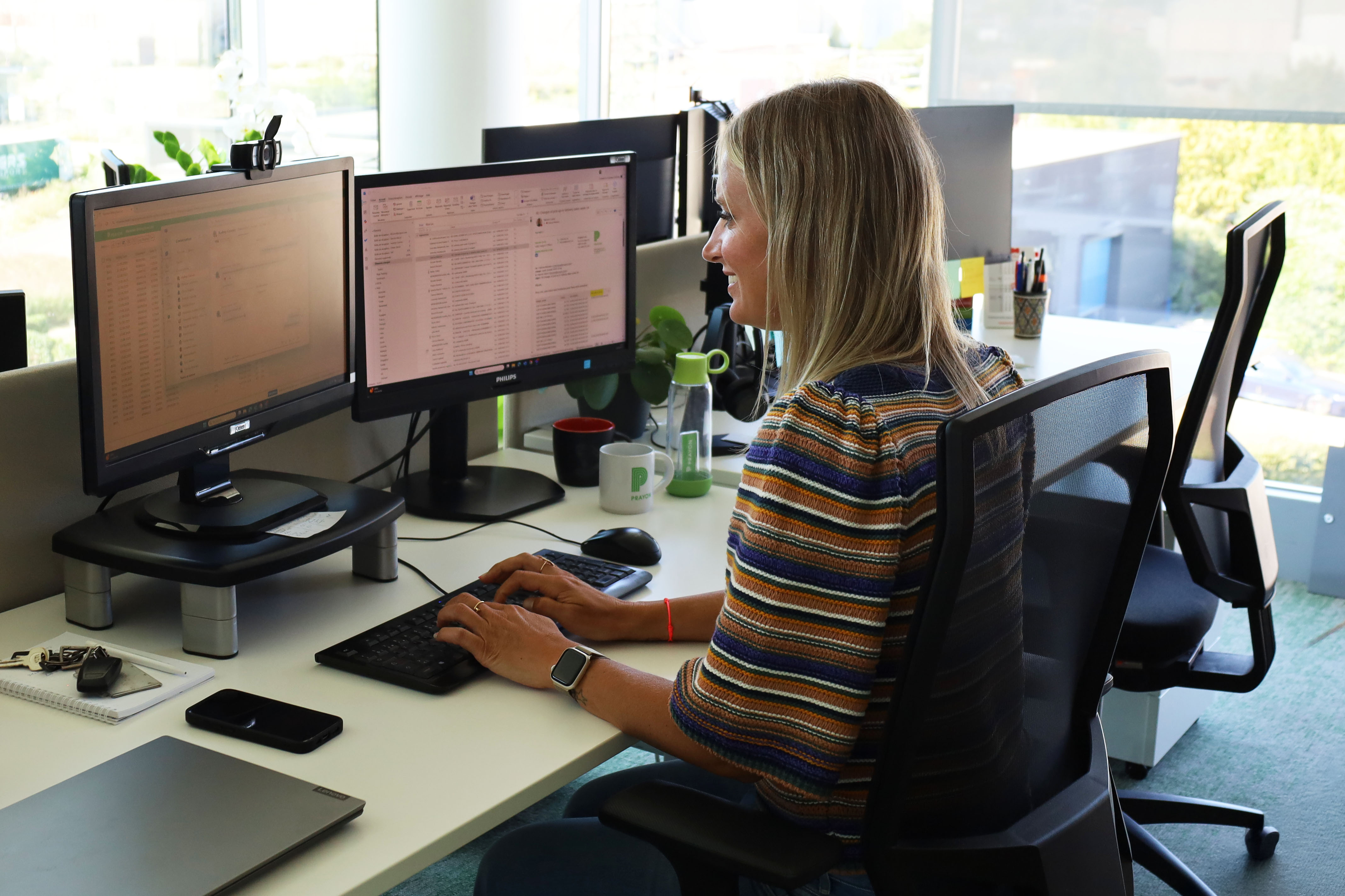 A blonde woman smiling sitting in front of two computer screens in an office environment 